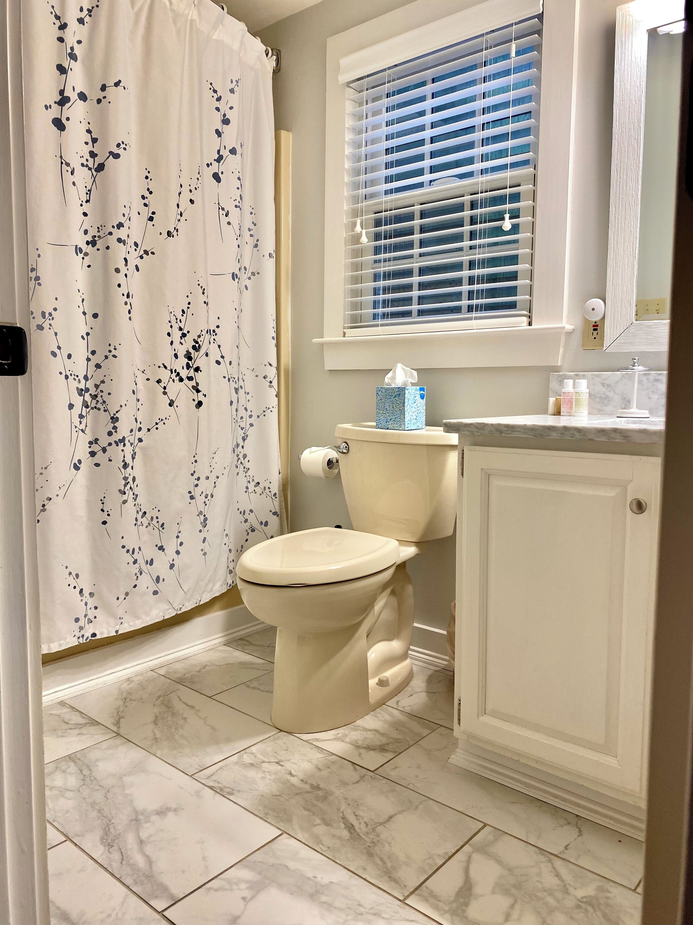 A bathroom with a toilet, a window with blinds, a shower curtain with a floral pattern, and a marble-tiled floor.