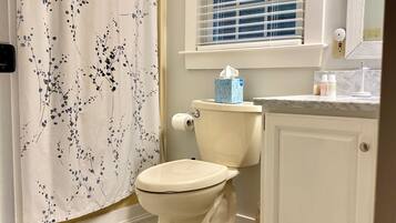A bathroom with a toilet, a window with blinds, a shower curtain with a floral pattern, and a marble-tiled floor.