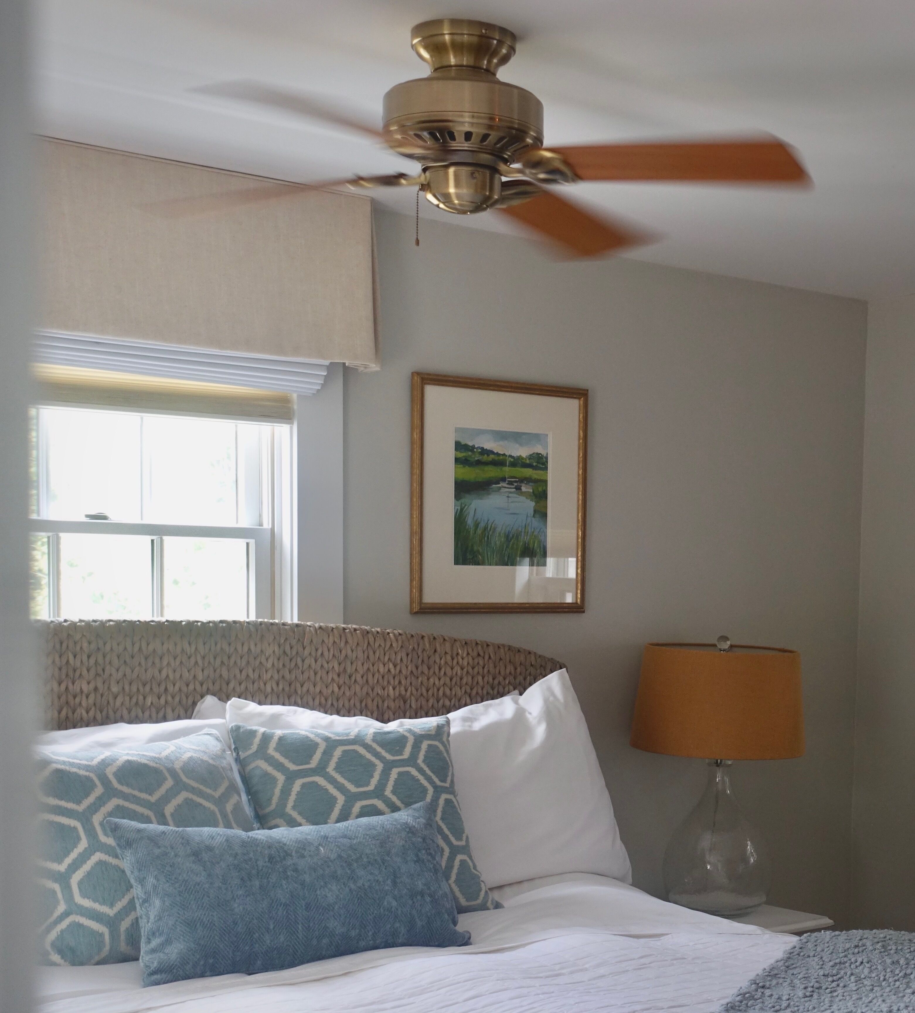 Une chambre à coucher avec un lit, un ventilateur de plafond, une fenêtre avec des stores, un tableau encadré au mur et une lampe de chevet.