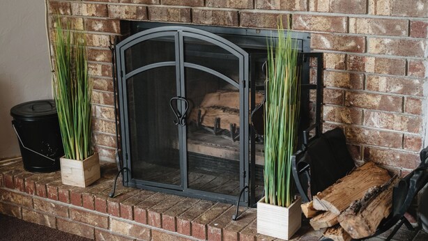 A brick fireplace with a metal screen, two potted plants, a black trash bin, and a stack of firewood. - Near Razorback Stadium & Attractions, Firepit (Springdale)
