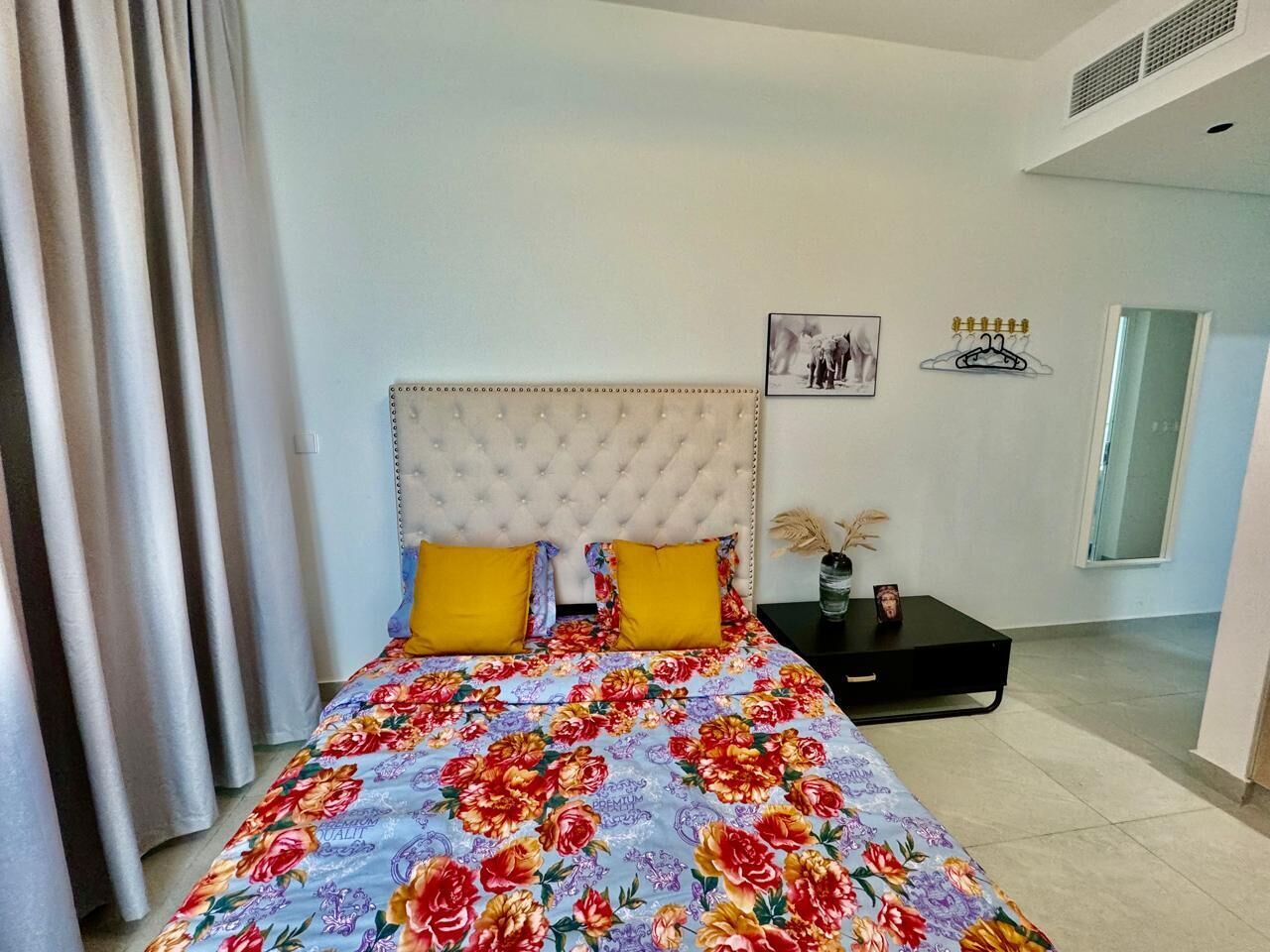 Ein Schlafzimmer mit einer gepolsterten Kopfteile, einer blumigen Bettdecke, zwei gelben Kissen, einem Nachttisch mit einer Vase und einem Bilderrahmen sowie einem Spiegel an der Wand.