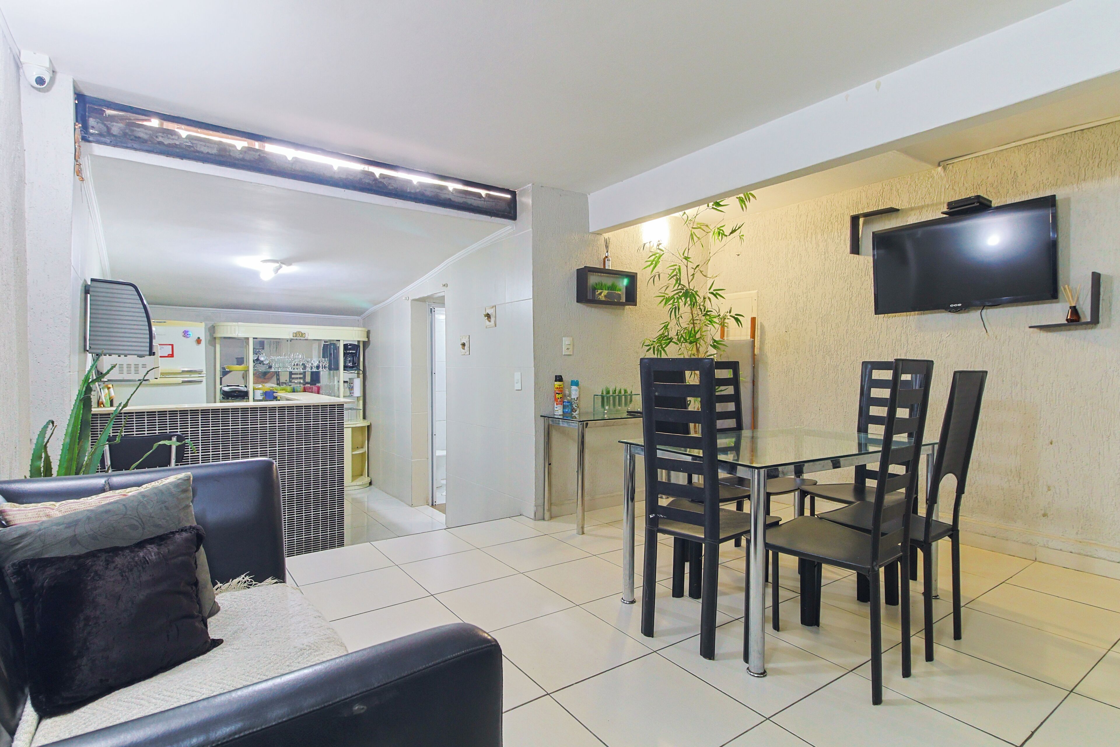 A dining area with a glass table and black chairs, a flat-screen TV mounted on the wall, and a kitchen in the background.