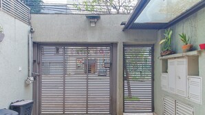 Una entrada con reja metálica, un cubo de basura pequeño y una planta en maceta sobre un estante.