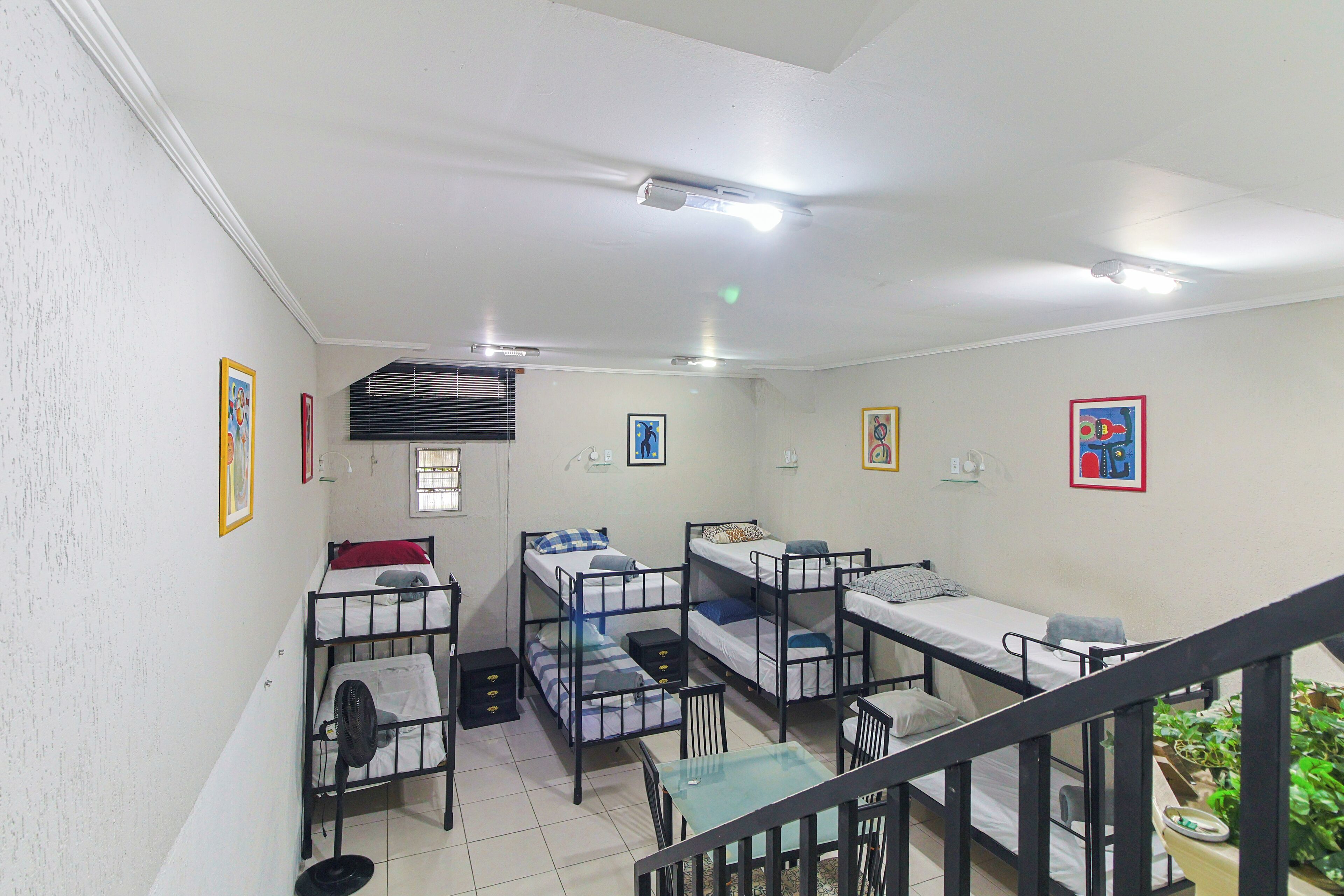 A dormitory room with bunk beds, a small table, and chairs. There are framed pictures on the walls and a ceiling fan.