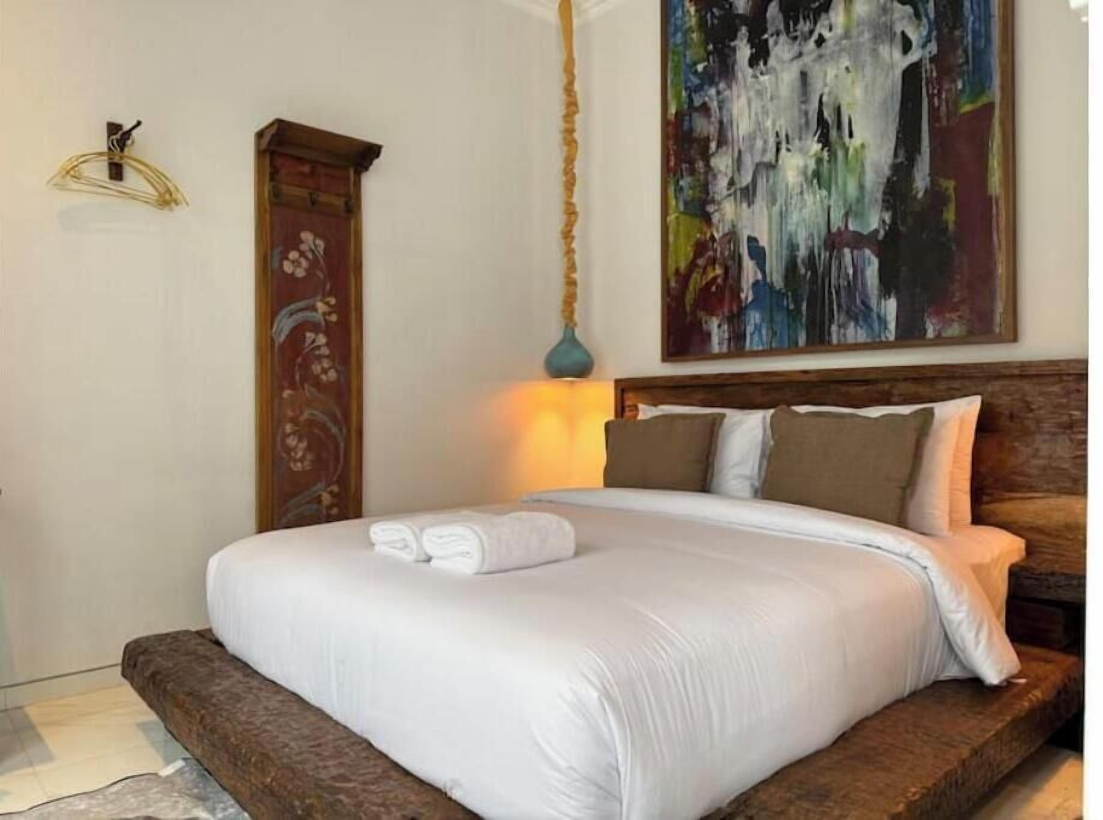 A neatly made bed with white linens and a brown headboard, a wooden cabinet with floral patterns, and a large abstract painting on the wall.