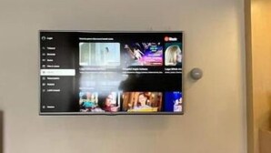 A flat-screen TV mounted on a wall above a wooden console table with a potted plant and a white object on it. - The Pines Smart Homes (Batu)