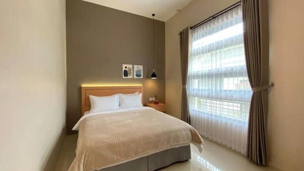 A hotel room with a bed, a nightstand, a lamp, and a window with curtains. - The Pines Smart Homes (Batu)