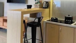 A modern kitchen with wooden cabinets, a black bar stool, a coffee maker, and pendant lights. - The Pines Smart Homes (Batu)