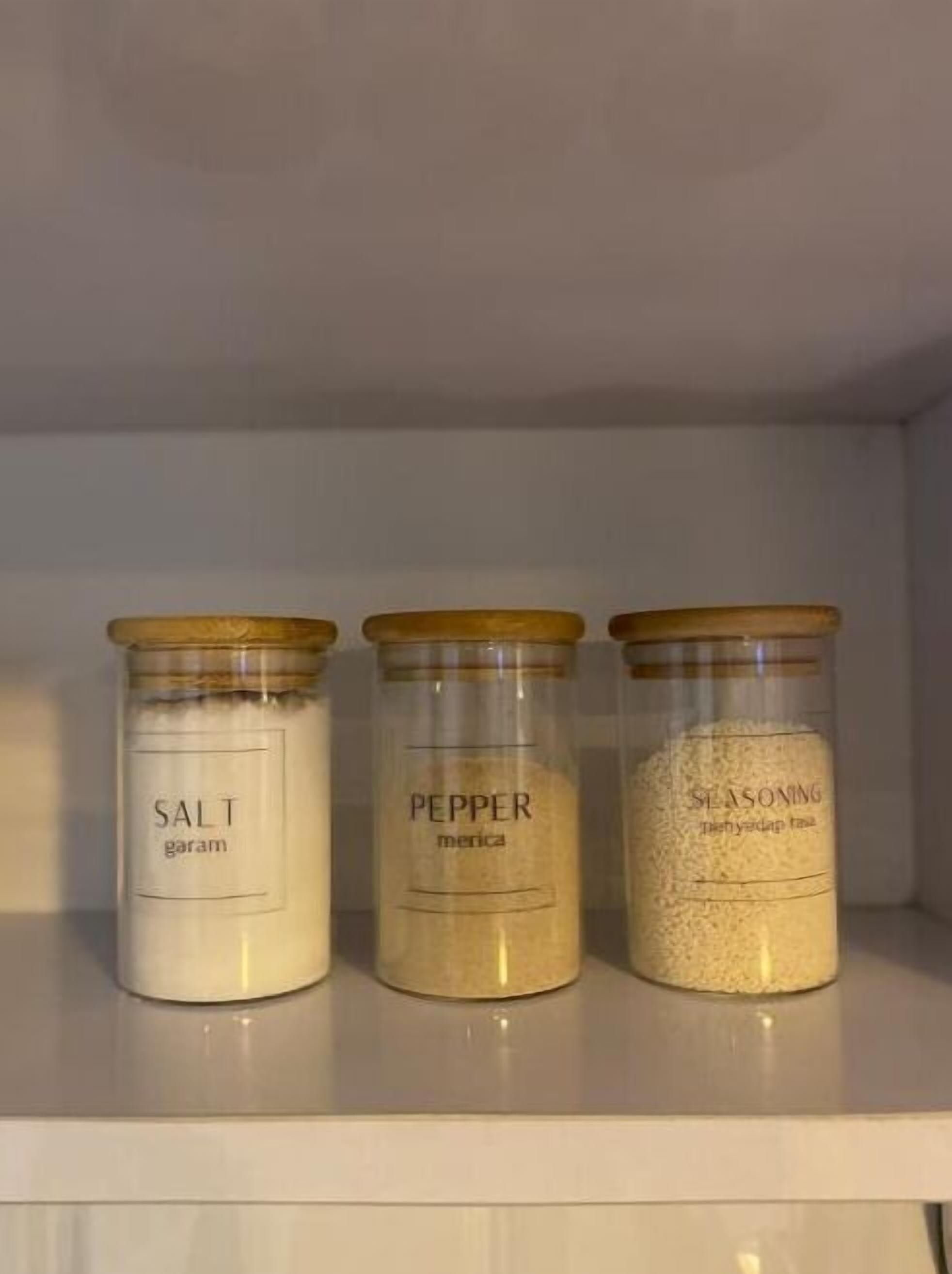Drei Gläser mit Holzböden auf einer weißen Ablagefläche. Die Gläser sind mit 'SALT', 'PEPPER' und 'SEASONING' beschriftet.