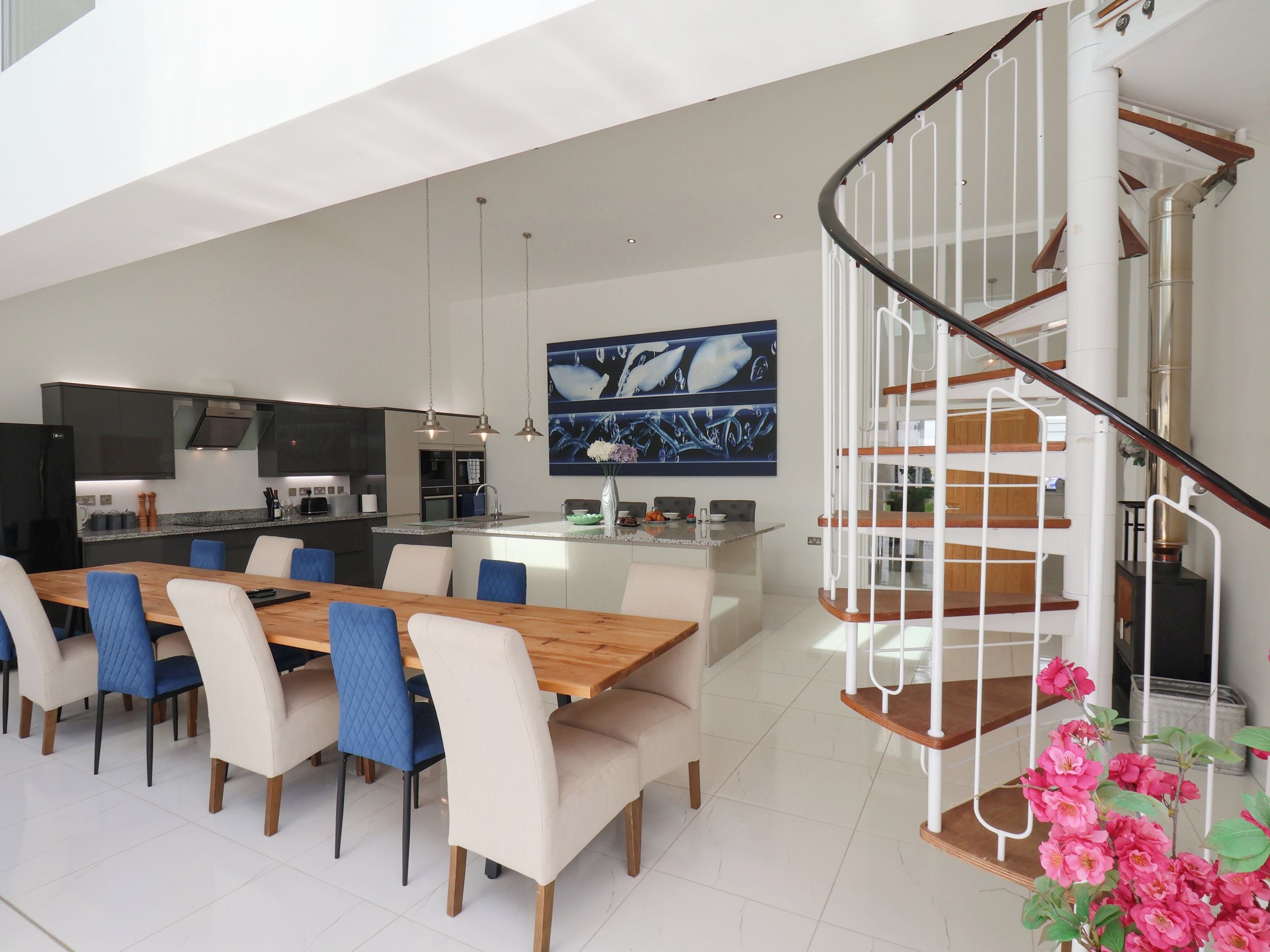 A modern kitchen with a central island, a dining area with a wooden table and chairs, and a spiral staircase.
