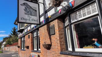 Eine Kneipe namens 'The Dog & Duck' mit einer traditionellen Backsteinfassade, großen Fenstern und einem hängenden Schild mit einer Hundeillustration.