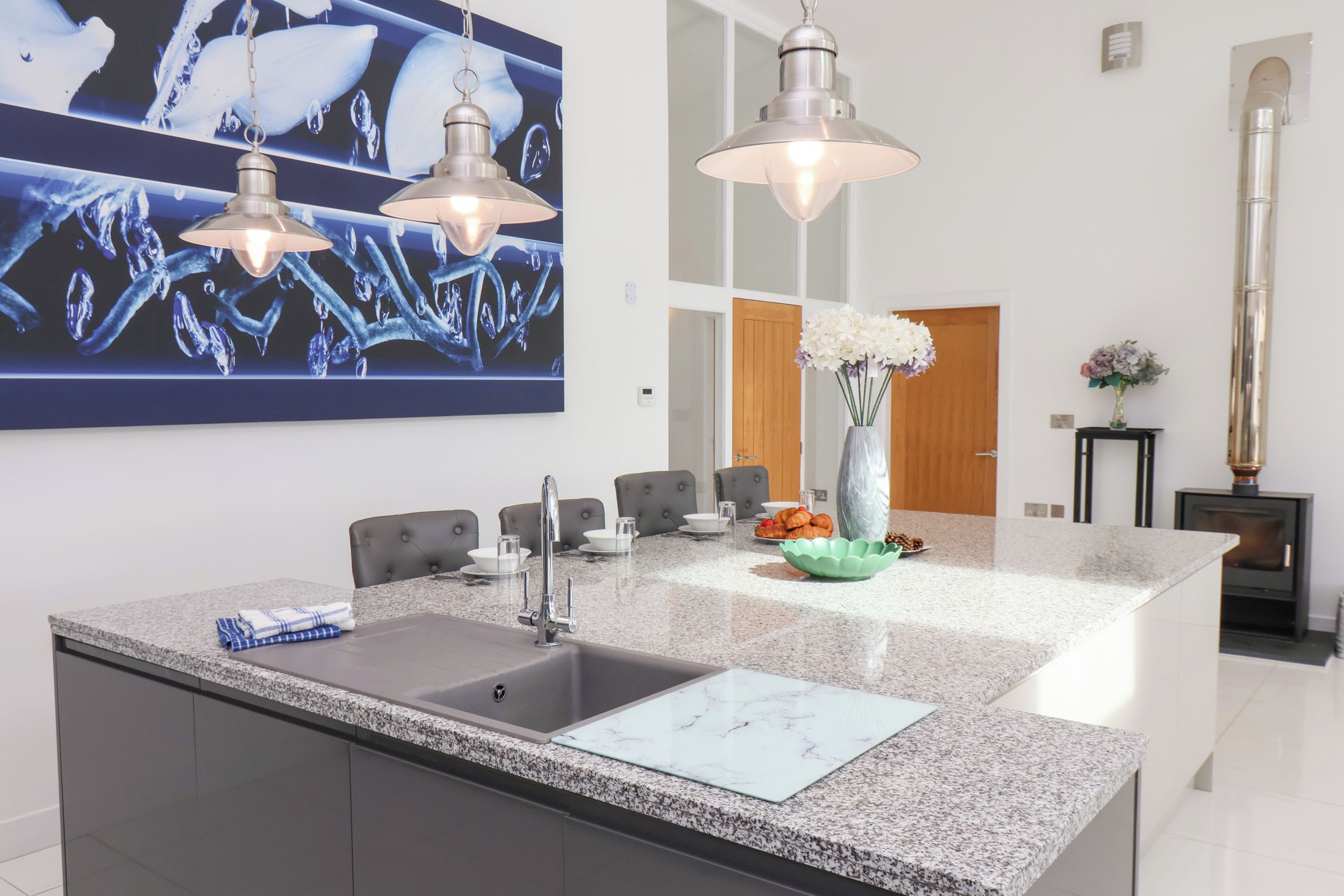 Una cocina moderna con encimera de granito, lavamanos de acero inoxidable y una isla grande. Hay tres lámparas colgantes, un jarrón con flores y un cuadro en la pared.