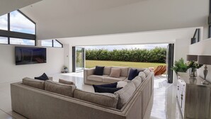 A modern living room with a large sectional sofa, a flat-screen TV mounted on the wall, and a view of a garden through an open door. - Meadow View (Beverley)