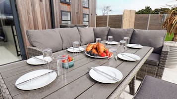 An outdoor dining area with a wooden table set for a meal, surrounded by grey cushioned seating.