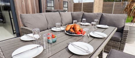 Un área de comedor al aire libre con una mesa de madera lista para una comida, rodeada de asientos acolchados de color gris.