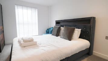 Una cama bien hecha con sábanas blancas y varias almohadas, un cabecero oscuro y una ventana con persianas.