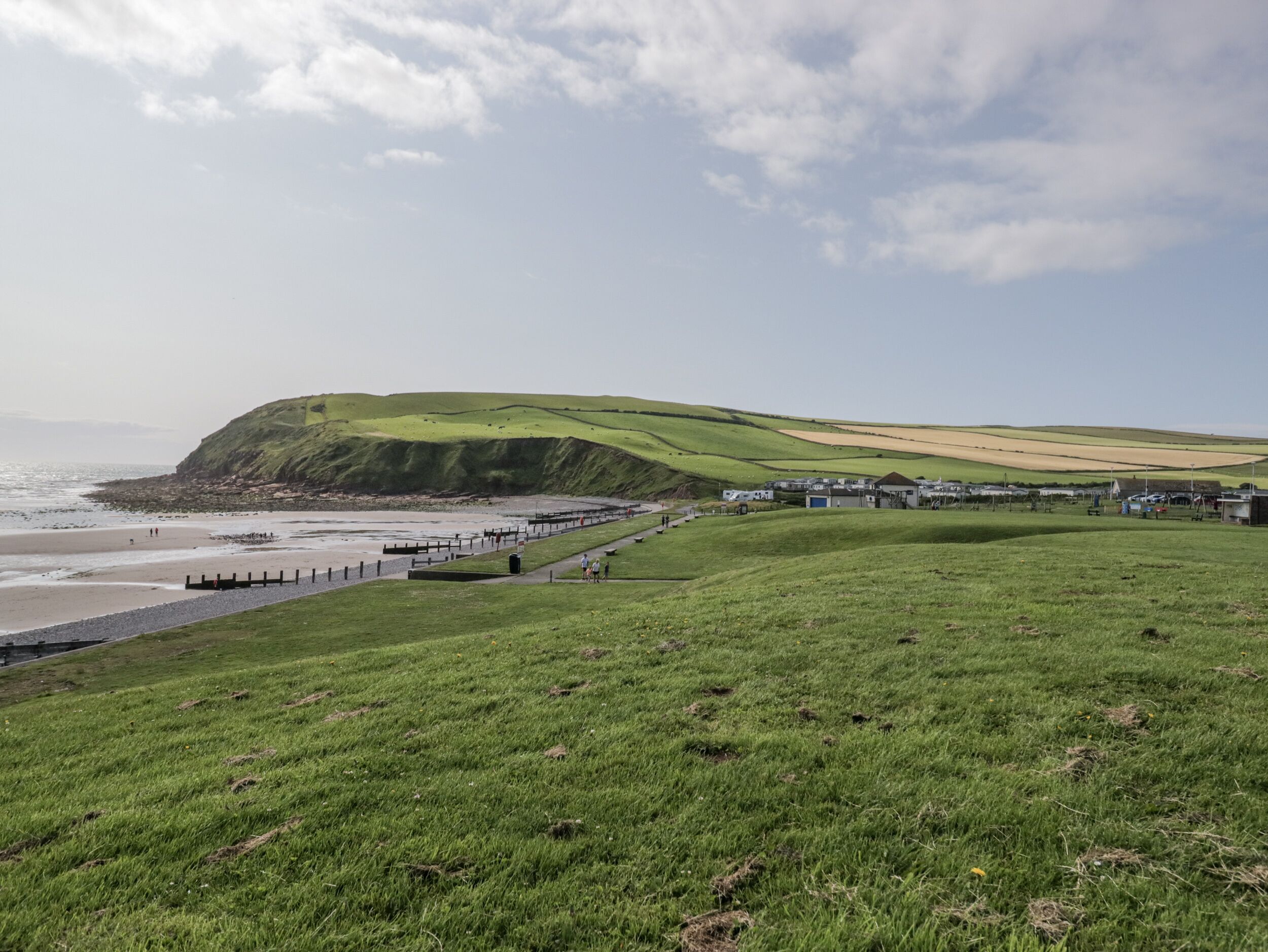 A coastal landscape with a grassy hill, a beach, and a row of buildings along the shore.