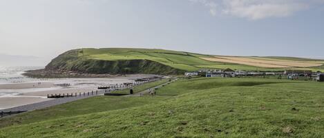 A coastal landscape with a grassy hill, a beach, and a row of buildings along the shore.
