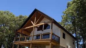 A two-story house with a wooden deck and a brick foundation, surrounded by trees.