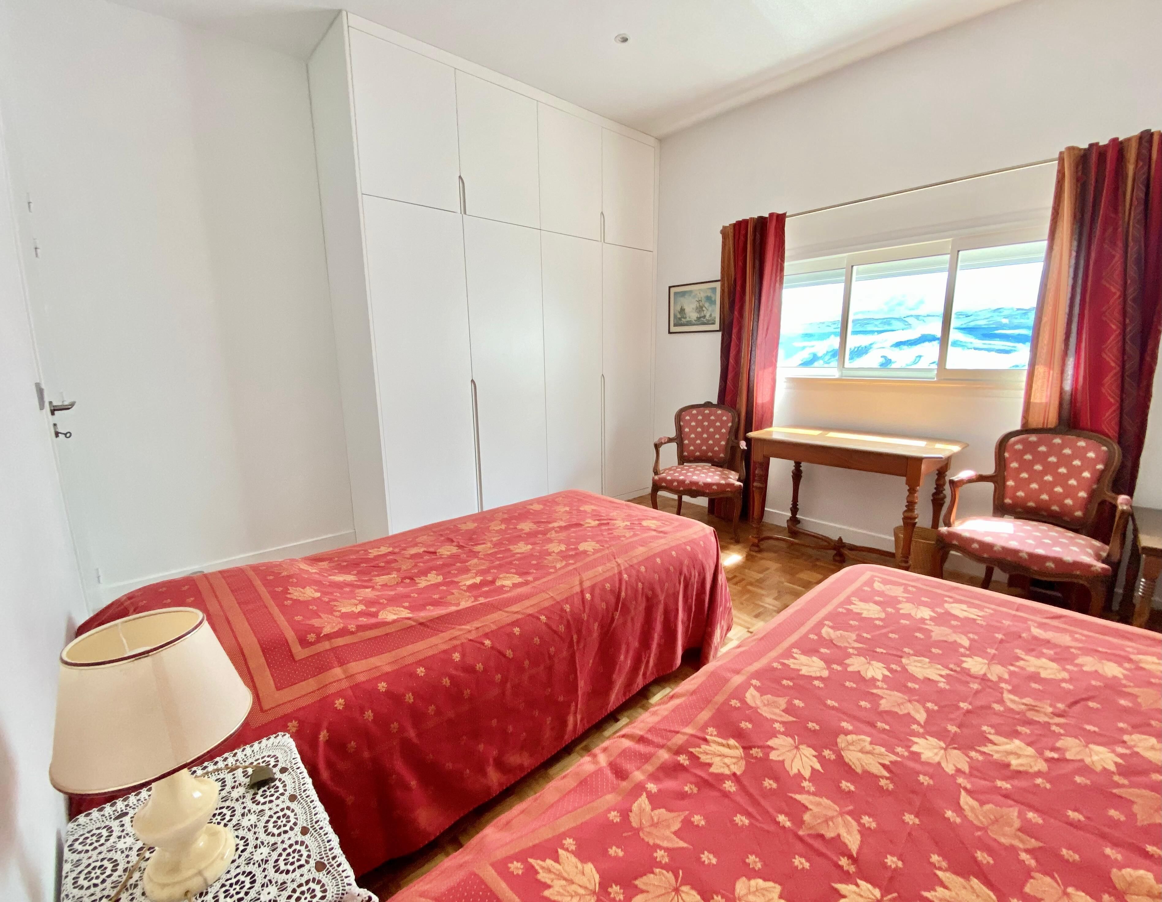 A room with two beds, a desk, and a chair. There is a window with curtains and a painting on the wall.
