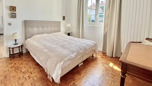 A bedroom with a large bed, a nightstand with a lamp, a window with curtains, and a view of buildings outside. - House Apartment whole garden rai (Biarritz)