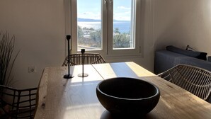 A dining area with a wooden table, two chairs, a black bowl, and a view of a coastal landscape through a window with a striped curtain. - Helios Thalassa 82 sqm (Tinos)