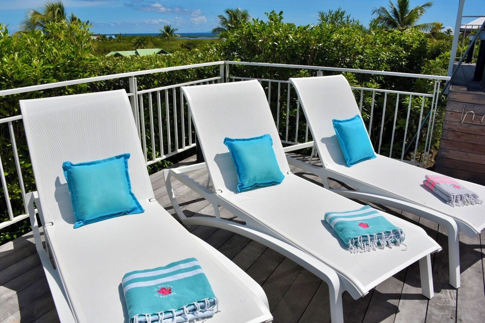 Three white lounge chairs with blue cushions and towels on a wooden deck with a white railing, overlooking a tropical landscape.