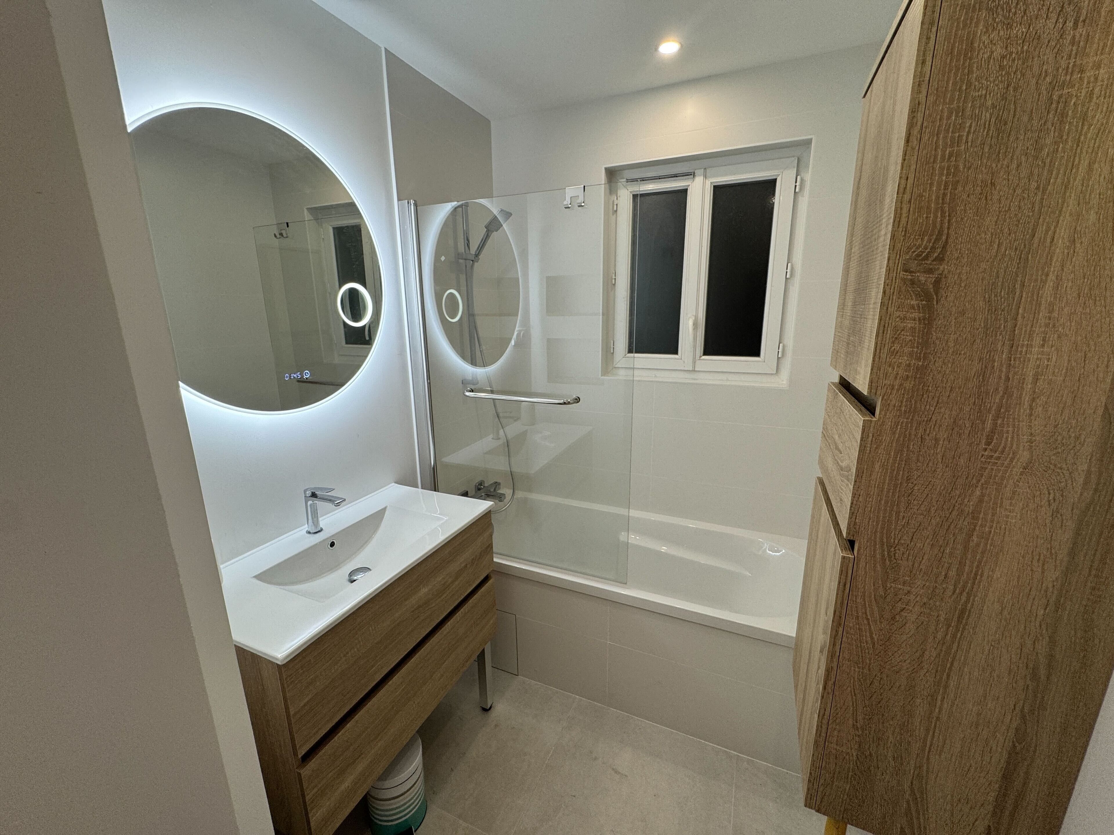 A modern bathroom with a glass-enclosed shower, a large mirror with circular lights, and a wooden vanity.
