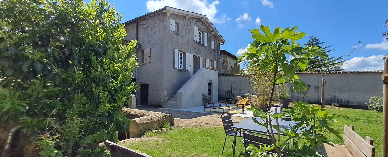 A rustic two-story house with a stone facade, a small front porch, and a backyard with a table and chairs.
