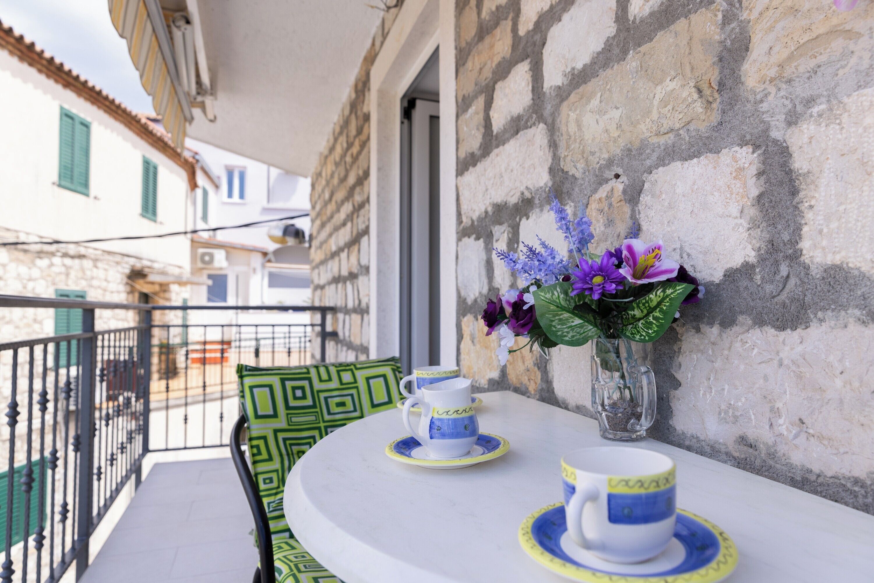 A balcony with a table set for two, a vase with purple flowers, and a stone wall.