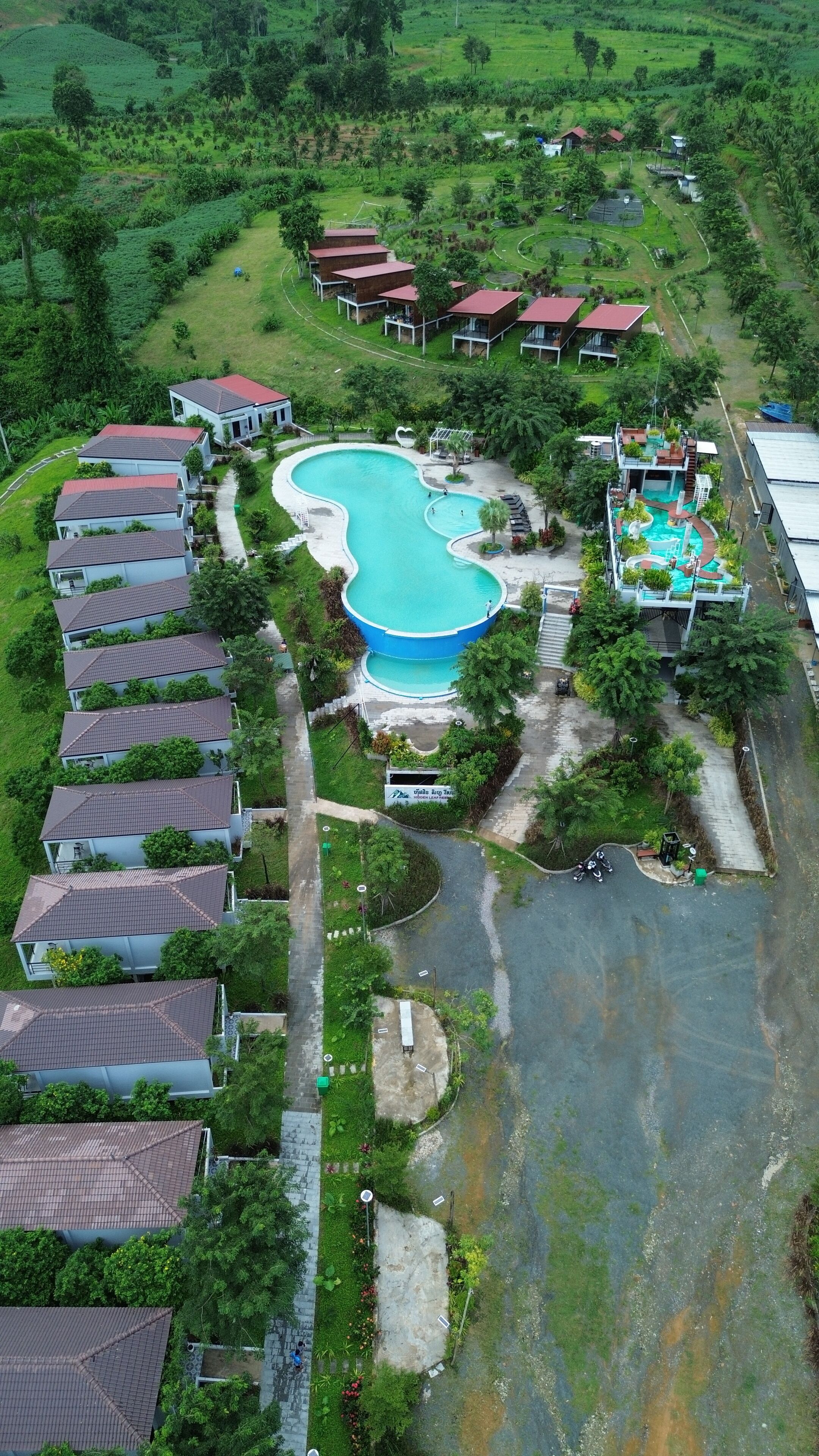 Vista aérea de una zona residencial con una gran piscina, varias casas con techos rojos y abundante vegetación alrededor de la propiedad.