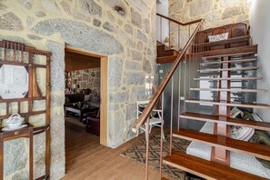 Interior de paredes de piedra con una escalera de madera, baranda de cristal y vista a una sala de estar.