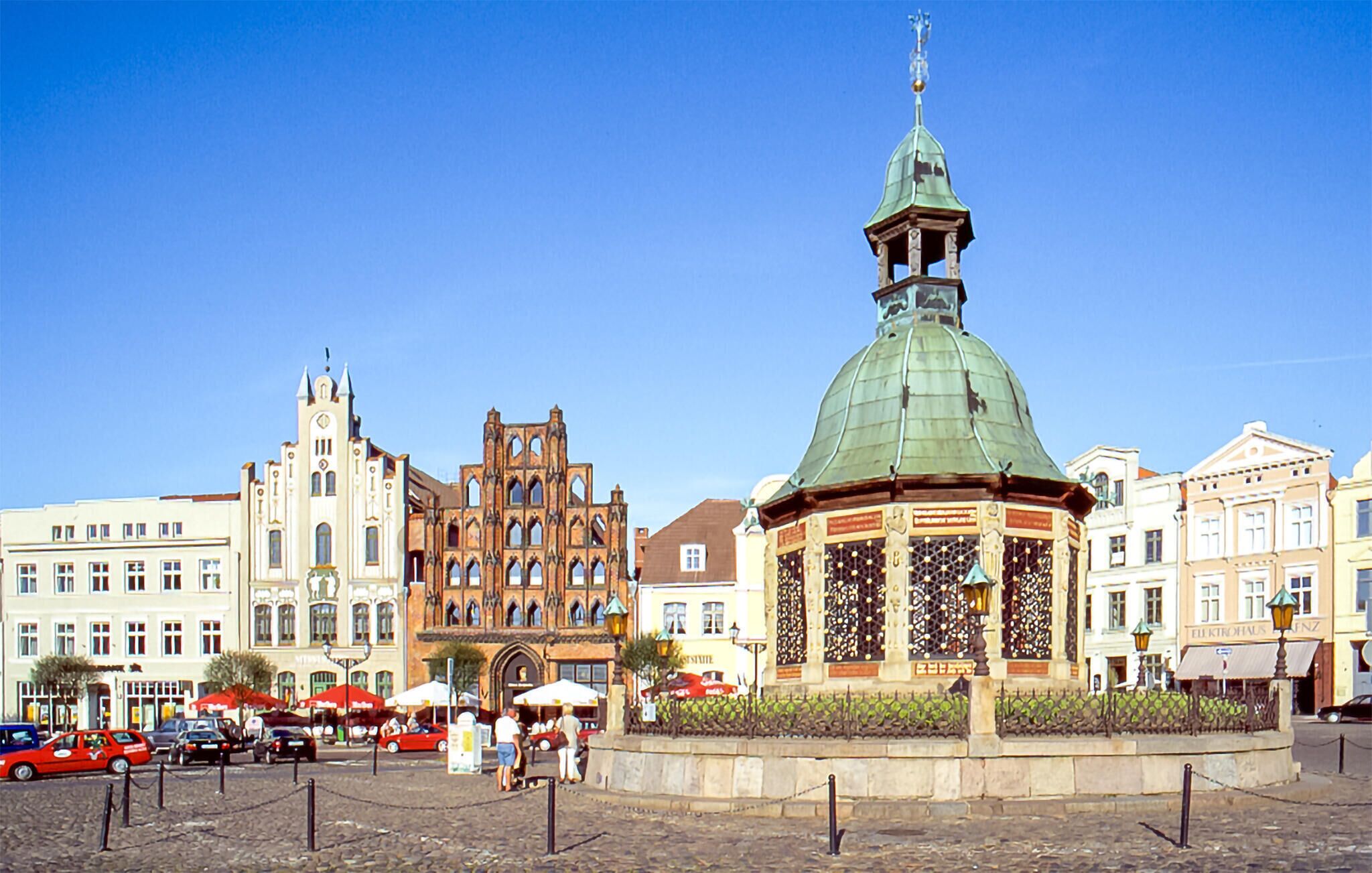 Ein historischer Platz mit einem markanten, grün-kuppeligen Pavillon, einem Gebäude im gotischen Stil sowie verschiedenen Geschäften und Cafés.