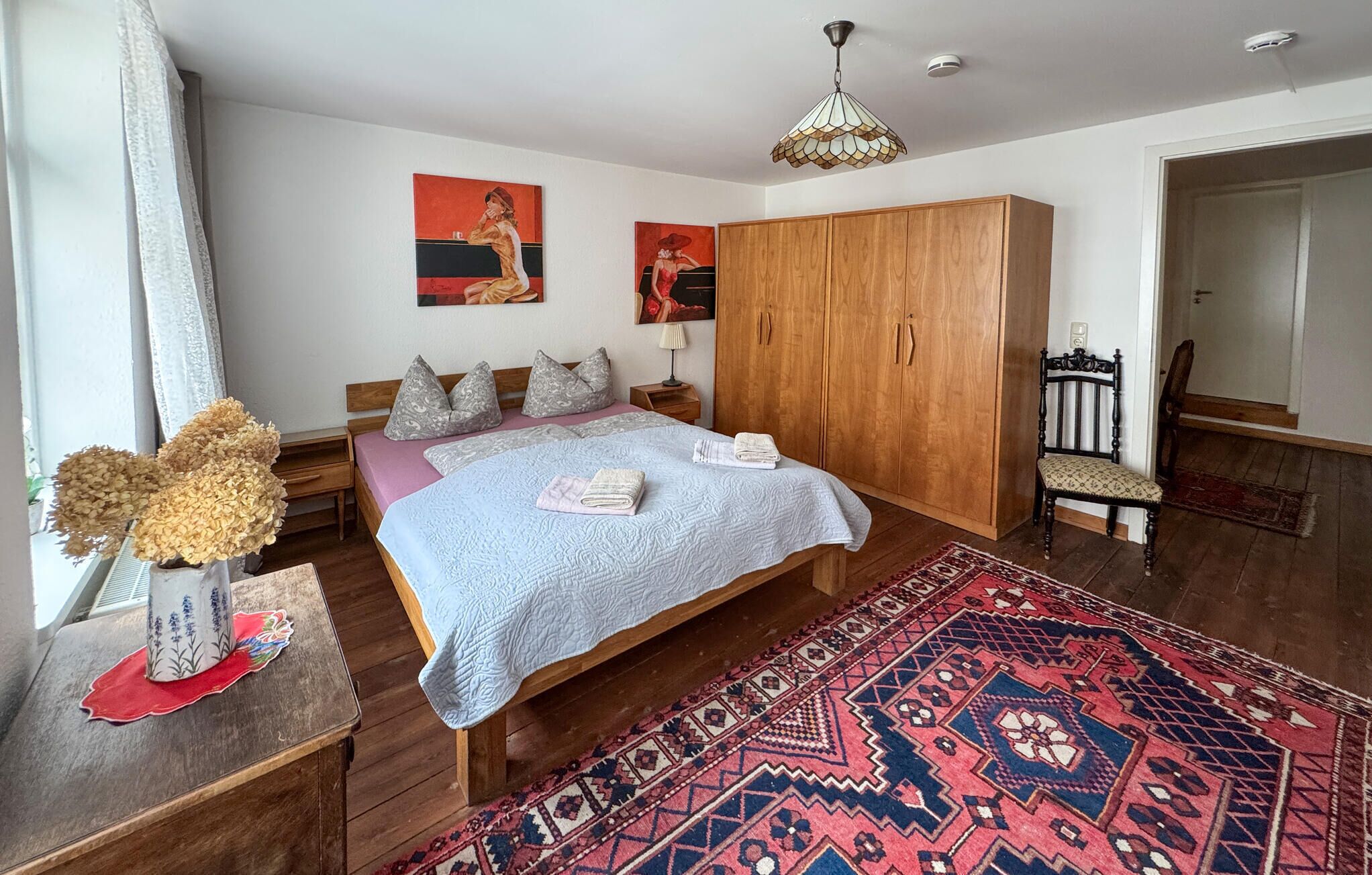 A bedroom with a large bed, wooden wardrobe, a chair, a rug, and artwork on the walls.