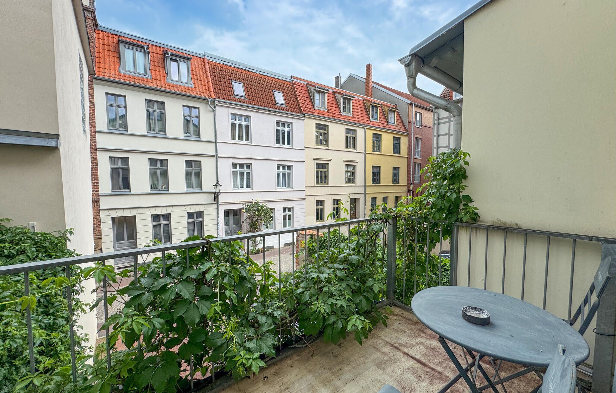 A balcony with a table and plants, overlooking a row of multi-story buildings with red-tiled roofs.