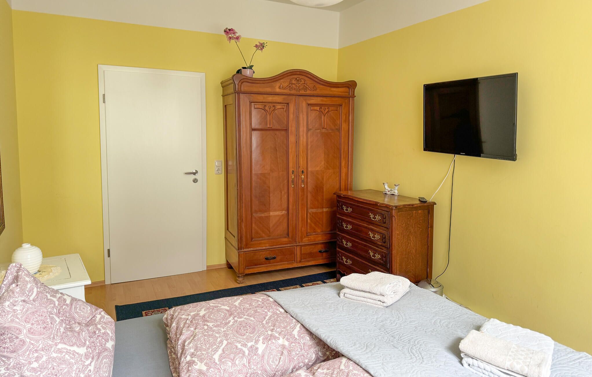 Um quarto com cama, guarda-roupa, penteadeira, televisão e porta.