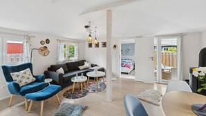 Ein großzügiger Wohnbereich mit einer Couch, Sesseln, einem Couchtisch und einem Esstisch. Ein großes Fenster mit Vorhängen, eine Tür zum Balkon und eine Wand mit Dekorationen.