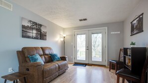 Ein Wohnzimmer mit braunem Ledersofa, einem kleinen Holztischchen, einem Bücherregal und einer zur Außenseite führenden Tür.