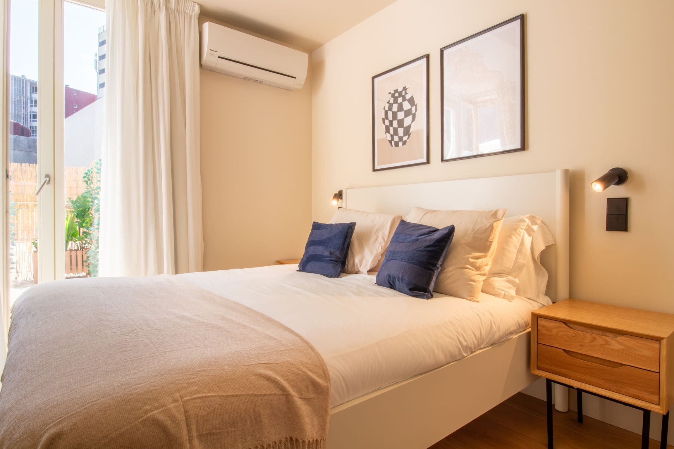 A hotel room with a bed, a nightstand, a drawer, an air conditioning unit, and two framed pictures on the wall.