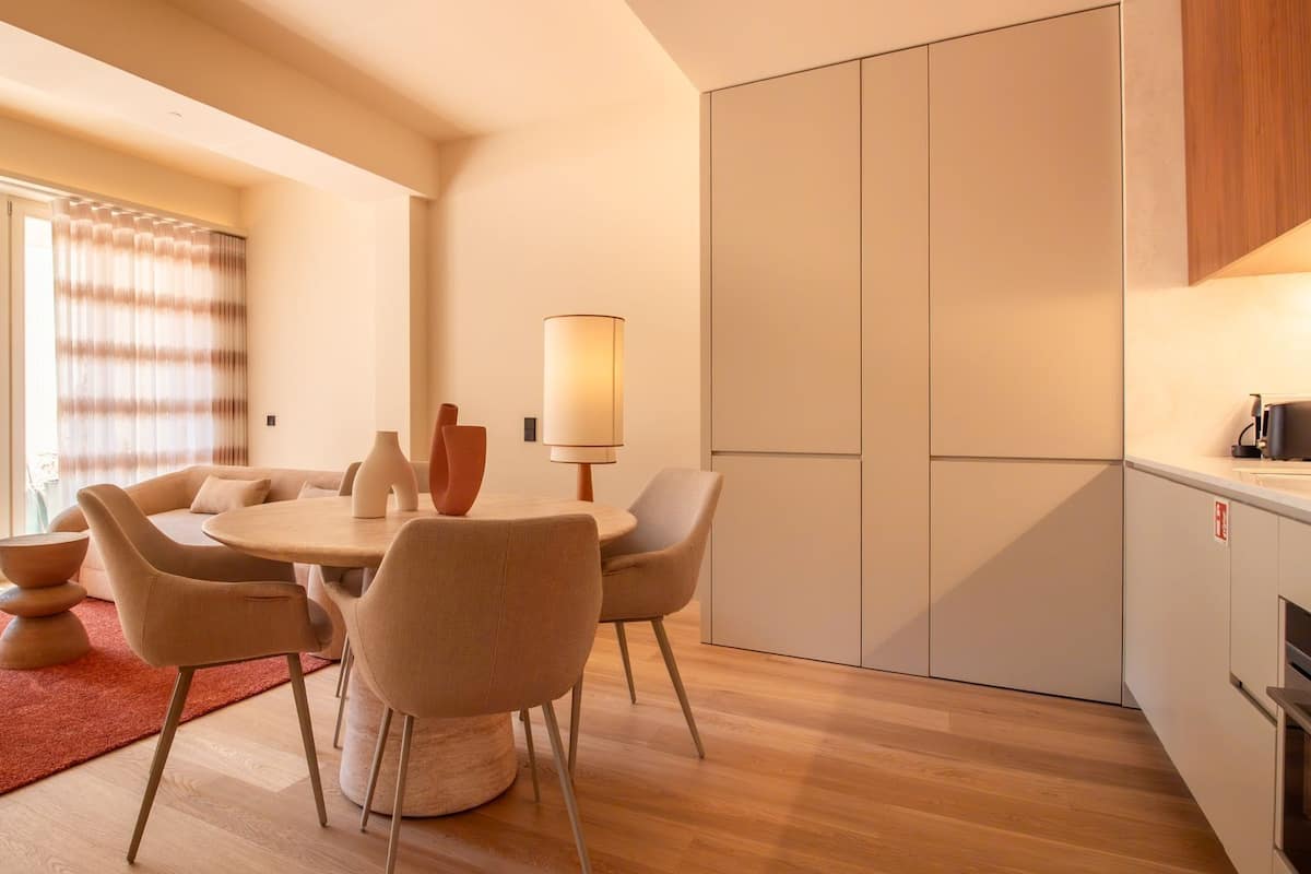 A modern living room with a round table, comfortable seating, a large window with curtains, and a kitchen area with cabinets.