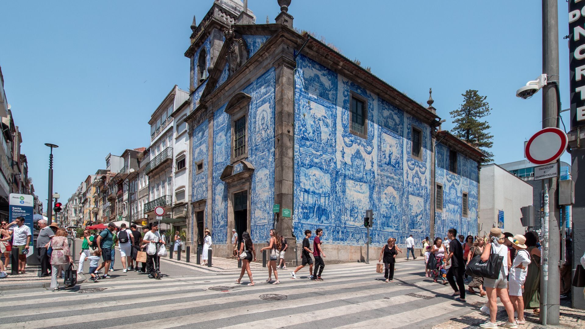 Une scène de rue avec un immeuble dont la façade est recouverte de carreaux bleus et blancs, un passage pour piétons et des personnes qui marchent.