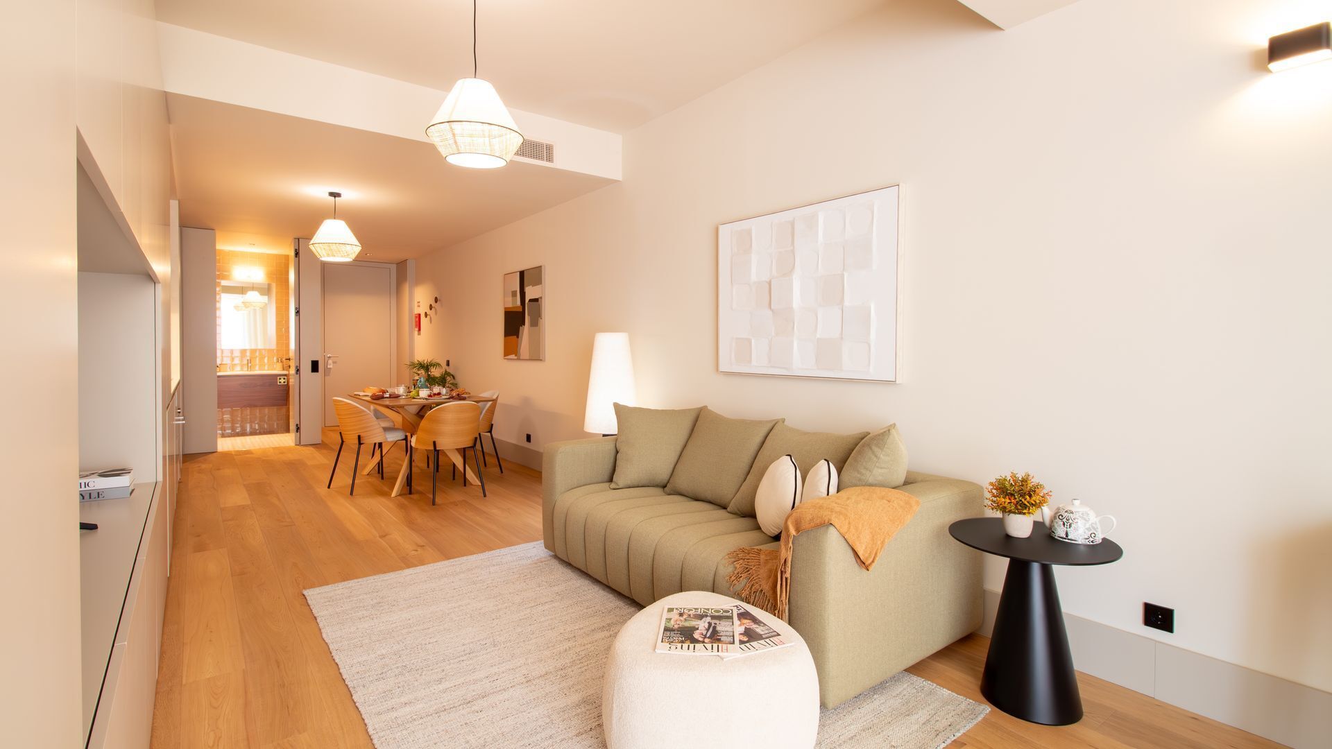 A modern living room with a sofa, dining table, and chairs. There is a coffee table and a lamp. The room has wooden flooring and white walls.