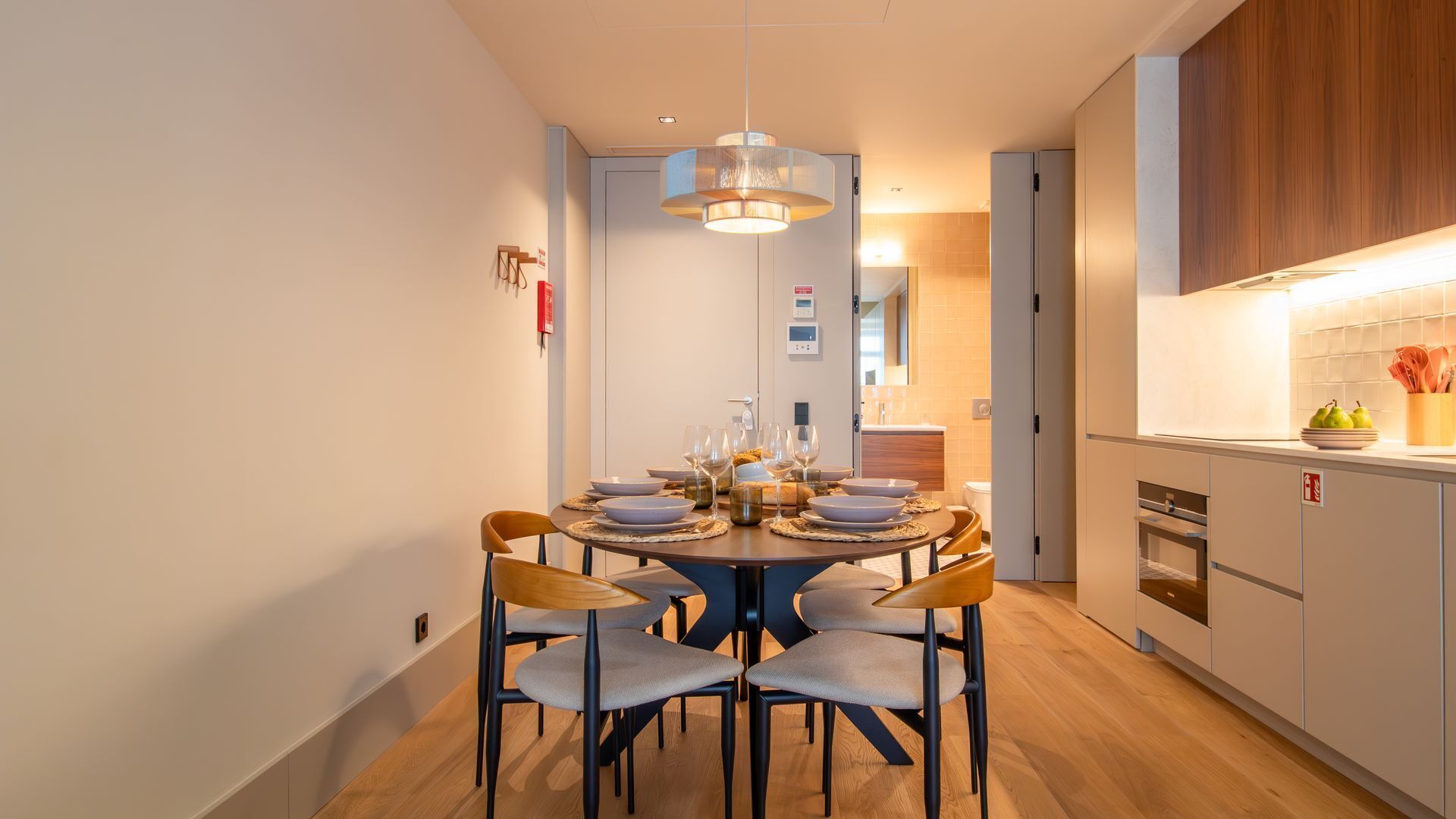 A modern dining area with a round table set for four, surrounded by chairs. A pendant light hangs above the table, and the room has a clean, minimalist design with wooden flooring and light-colored walls.