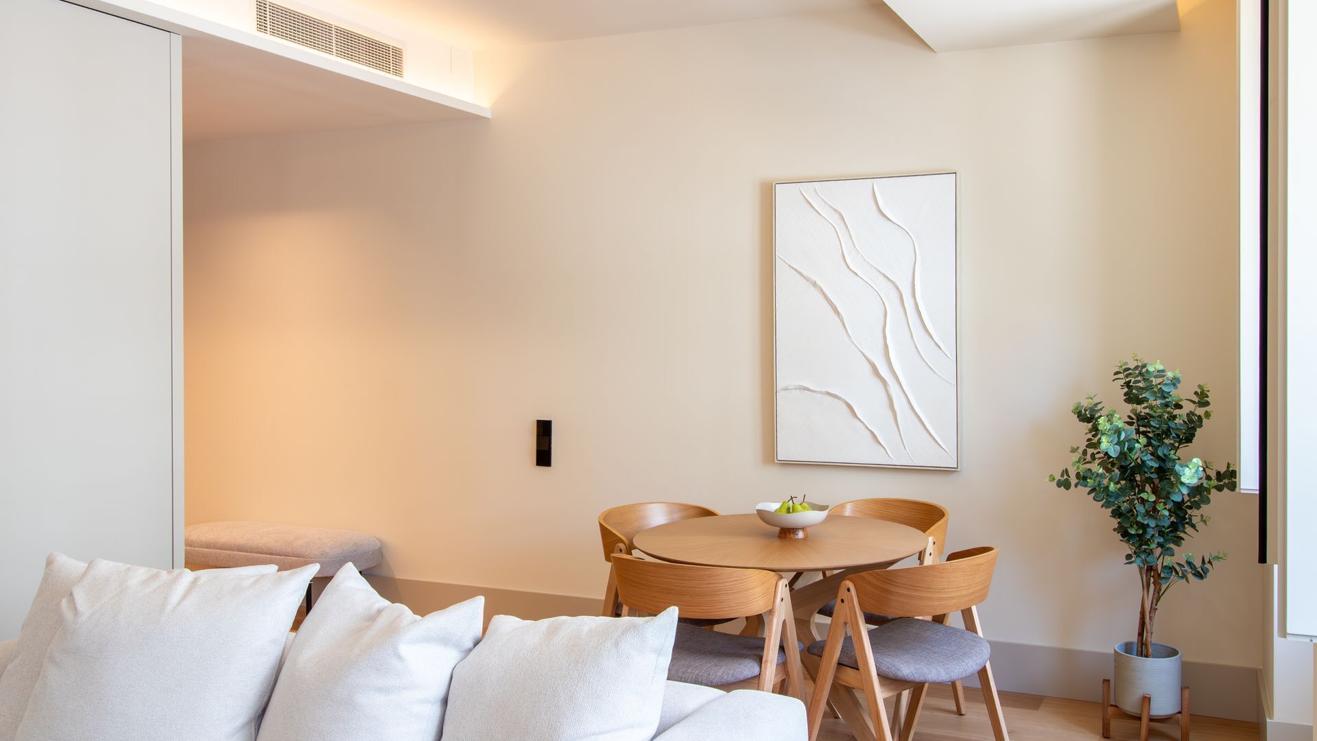 A modern living room with a sofa, a dining table with chairs, a wall-mounted abstract art piece, and a potted plant.
