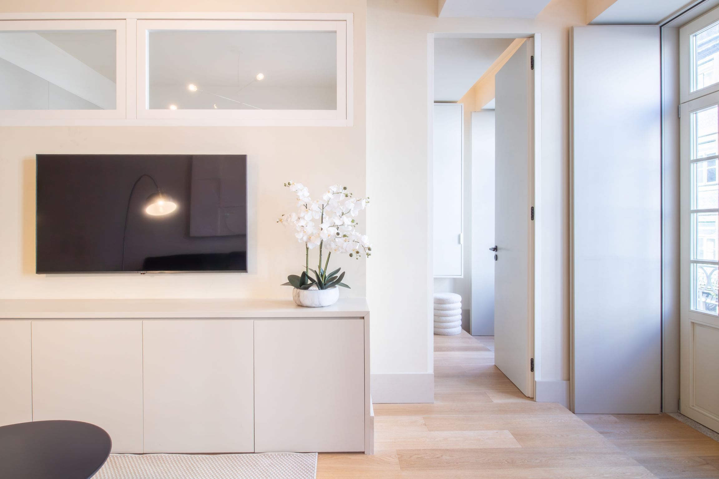 Interior moderno com televisão de tela plana presa a uma parede branca, uma orquídea em vaso sobre um console branco, e um corredor que leva a outro cômodo.