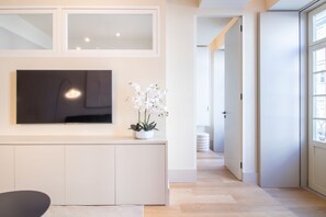 Un interior moderno con un televisor de pantalla plana montado en una pared blanca, una orquídea en maceta sobre una consola blanca y un pasillo que lleva a otra habitación.