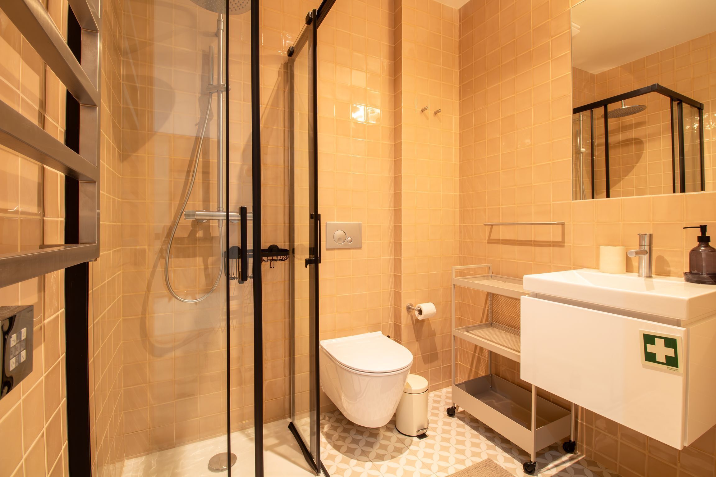 A modern bathroom with a glass shower enclosure, a toilet, and a sink with a green cross symbol.