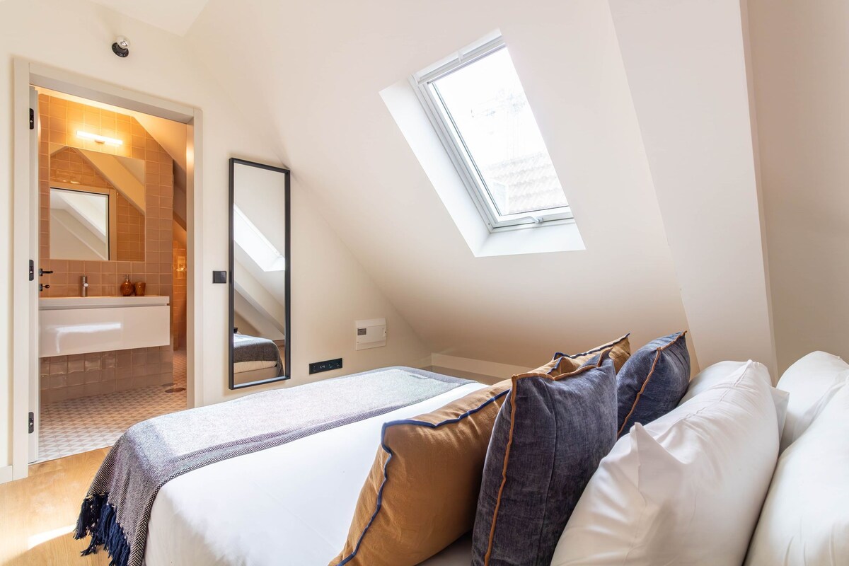 A bedroom with a skylight, a bed with pillows, a mirror, and a bathroom with a sink and mirror.