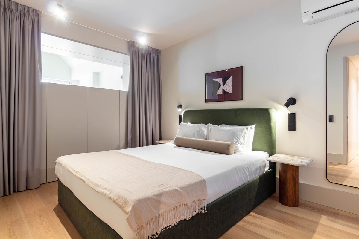 A modern hotel room with a large bed, a headboard, a bedside table, a mirror, and a window with curtains.
