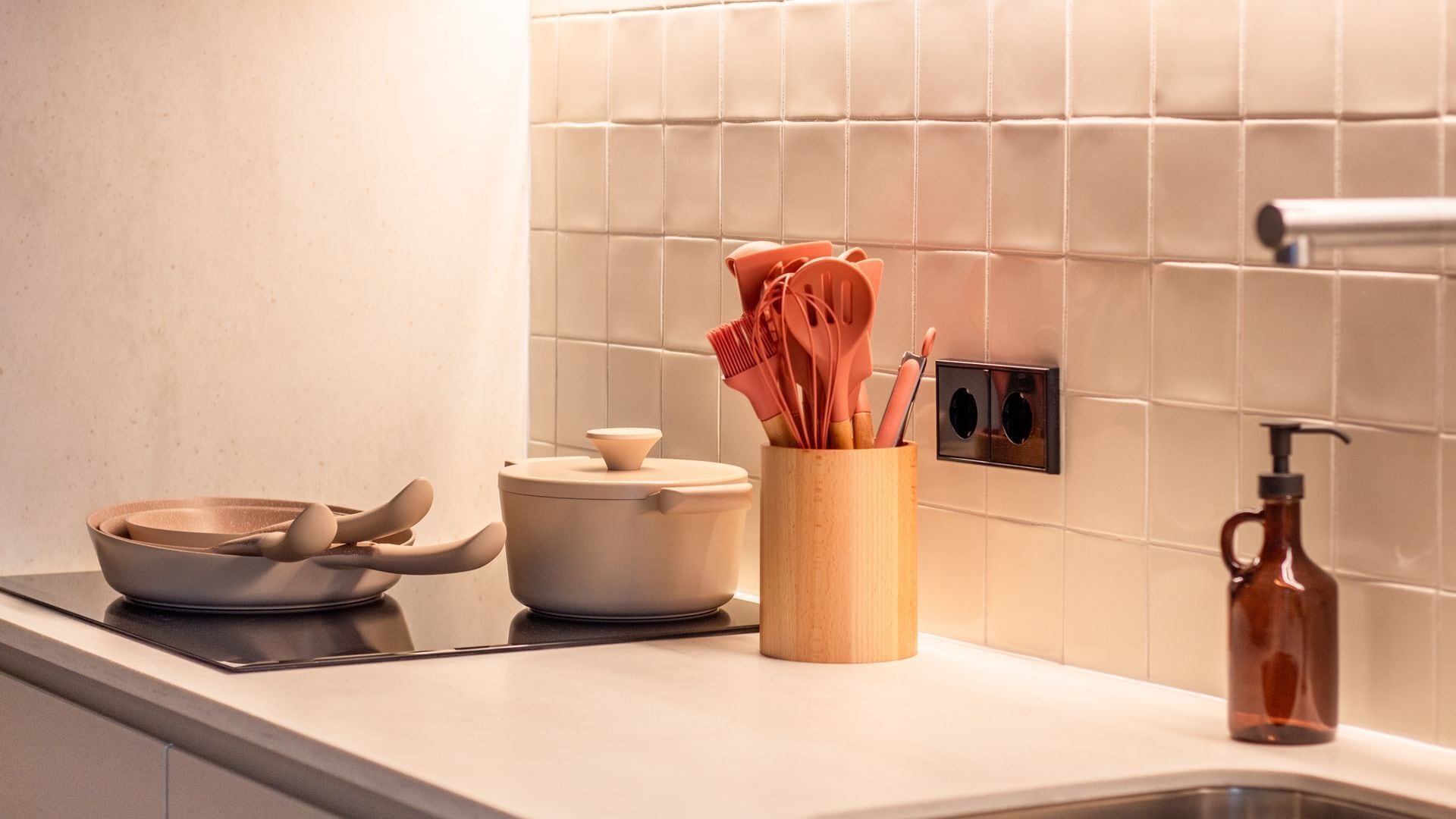 Una cocina con salpicadero de azulejos blancos, un utensilio de madera, una olla de acero inoxidable y un dispensador de jabón.